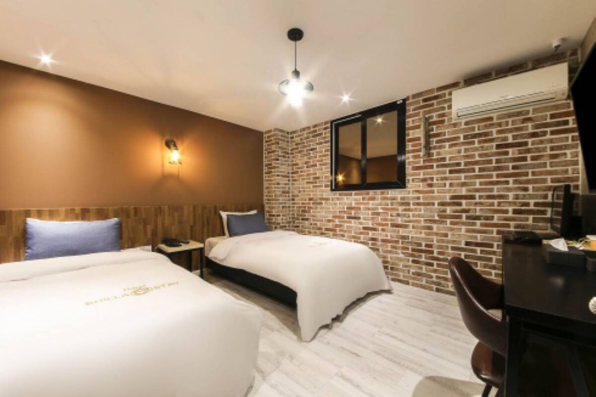 A hotel room with two beds, a desk, a chair, a brick wall, and an air conditioning unit.