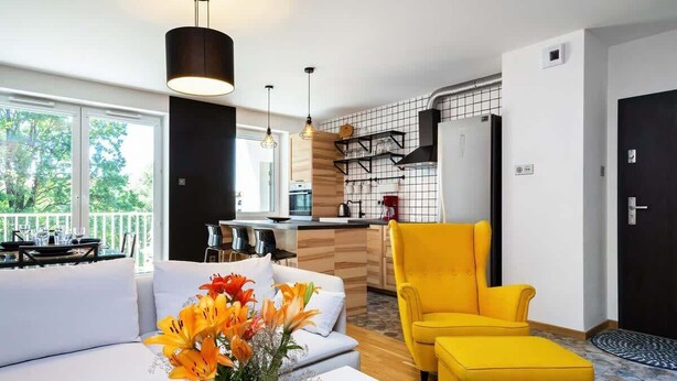 Una sala de estar moderna con un sofá, un sillón amarillo y una mesa de centro. Hay una zona de cocina con encimera y una lámpara colgante.