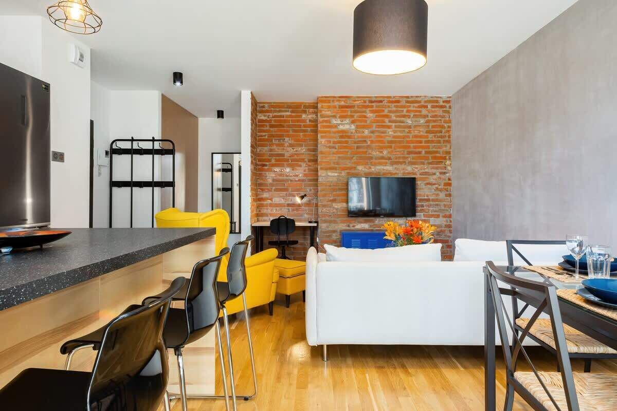 Eine moderne Küche mit einem Edelstahlkühlschrank, ein Essbereich mit einem Holztisch und Stühlen, ein weißes Sofa und eine Ziegelwand mit einem Flachbildfernseher.