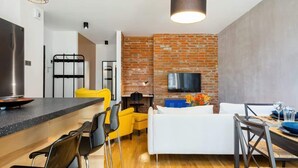 Una cocina moderna con un refrigerador de acero inoxidable, un comedor con una mesa y sillas de madera, un sofá blanco y una pared de ladrillo con un televisor de pantalla plana.