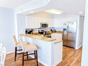 A modern kitchen with white cabinetry, a central island with bar stools, stainless steel appliances, and a wooden floor. - Ashworth 1406 (North Myrtle Beach)