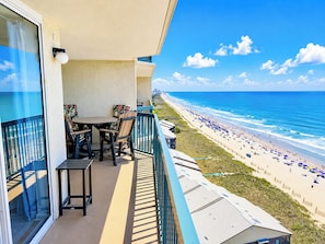 Ein Balkon mit Tisch und Stühlen, der Blick auf einen Strand mit Sonnenschirmen und Menschen fällt.