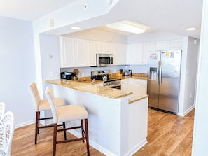 A kitchen with white cabinets, a stainless steel refrigerator, a microwave, and a granite countertop island with two bar stools.