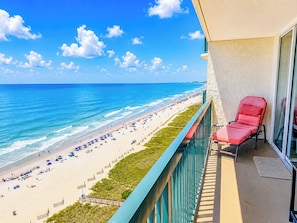 Ein Balkon mit einem roten Stuhl, der Blick auf einen Strand mit Sonnenschirmen und Menschen freigibt.
