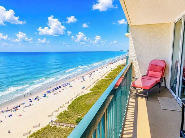A balcony with a red chair, overlooking a beach with umbrellas and people.