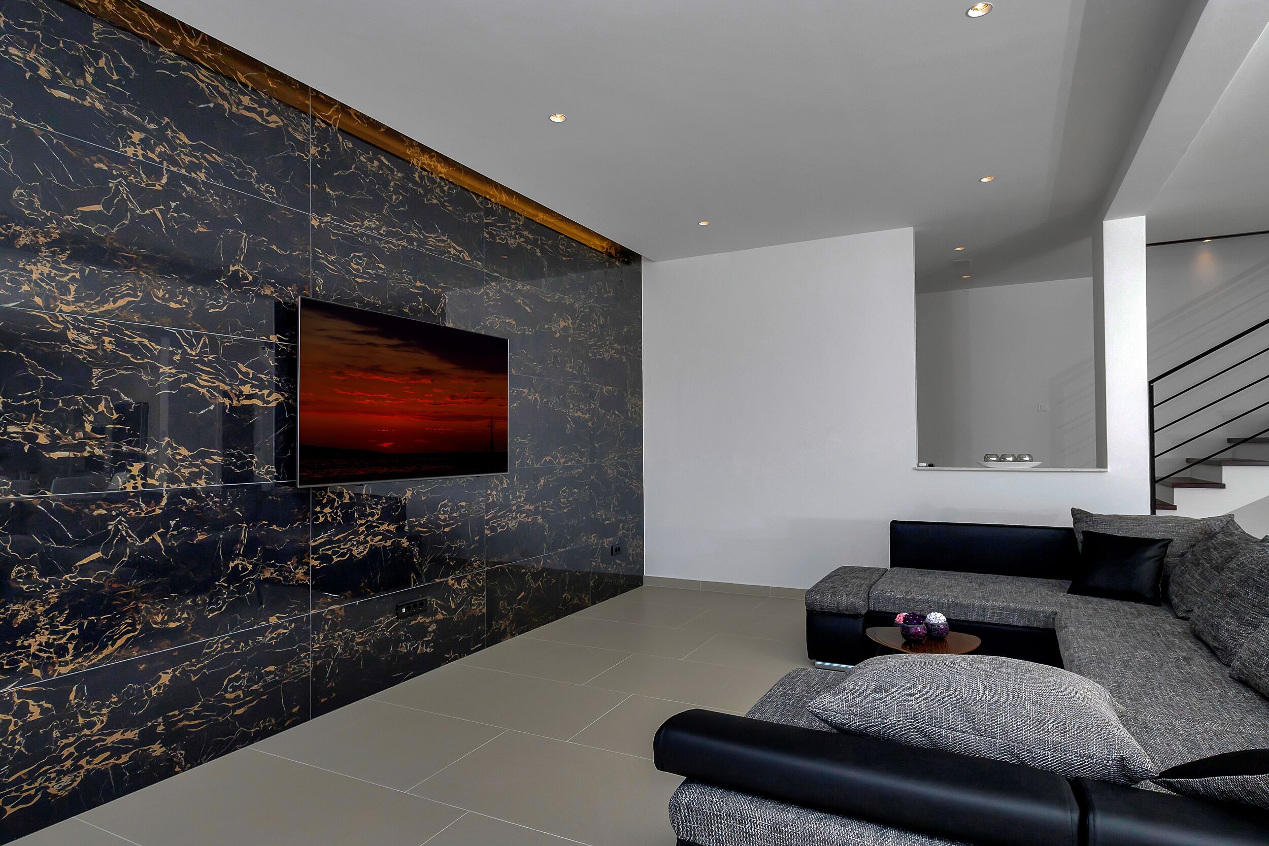 A modern living room with a large flat-screen TV mounted on a black marble wall, a grey sectional sofa, and a staircase in the background.