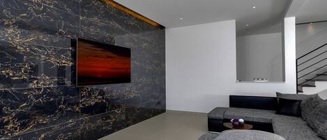 A modern living room with a large flat-screen TV mounted on a black marble wall, a grey sectional sofa, and a staircase in the background.