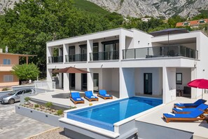 A modern two-story building with a swimming pool, outdoor seating, and a view of a mountainous landscape. - Modern holiday home with infinity pool, three bedrooms, terrace, sea view (Bast)