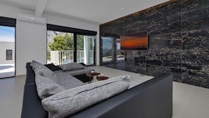 A modern living room with a large sectional sofa, a flat-screen TV mounted on a black marble wall, and a balcony with a view of the outdoors. - Modern holiday home with infinity pool, three bedrooms, terrace, sea view (Bast)