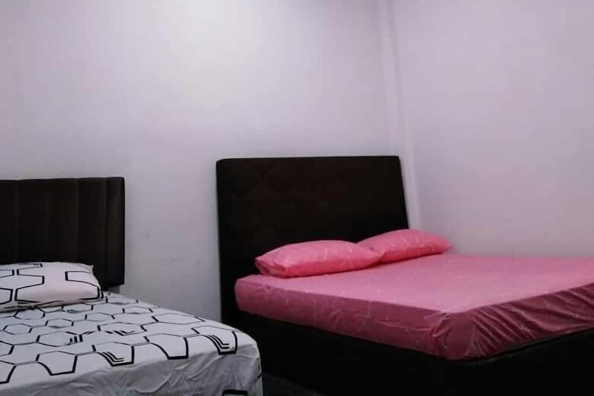 A room with two beds, one with a patterned bedspread and the other with pink bedding. There is a ceiling light fixture with three bulbs.