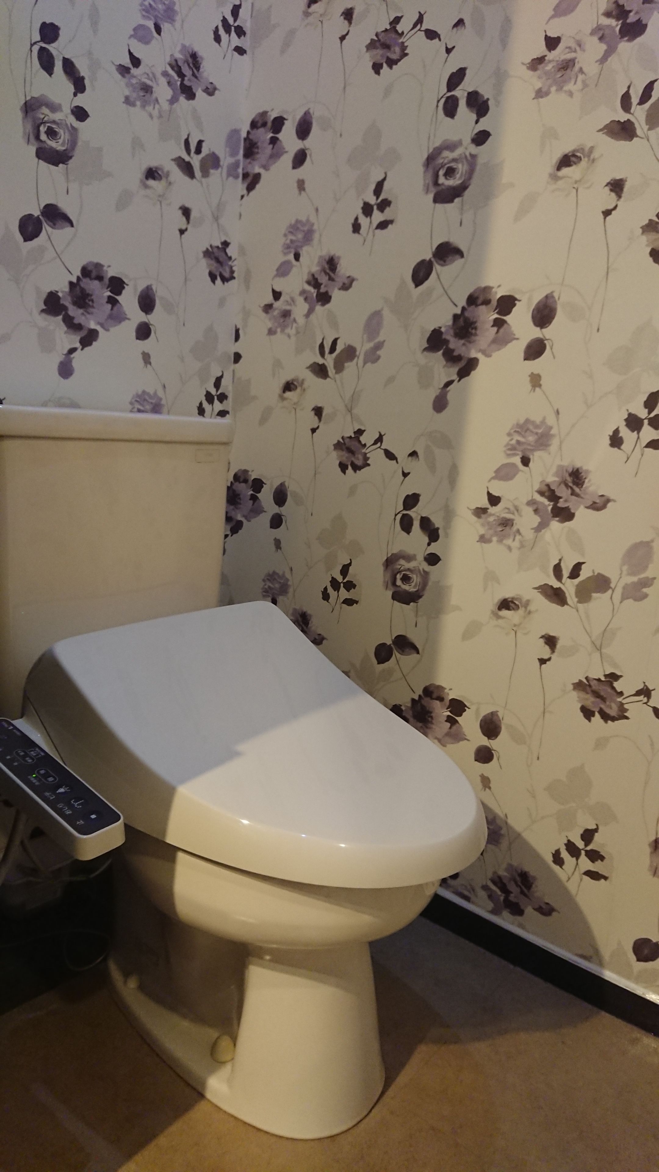 Des toilettes dotées d’un panneau de commande, d’un papier peint à motifs floraux et d’un sol de couleur beige.