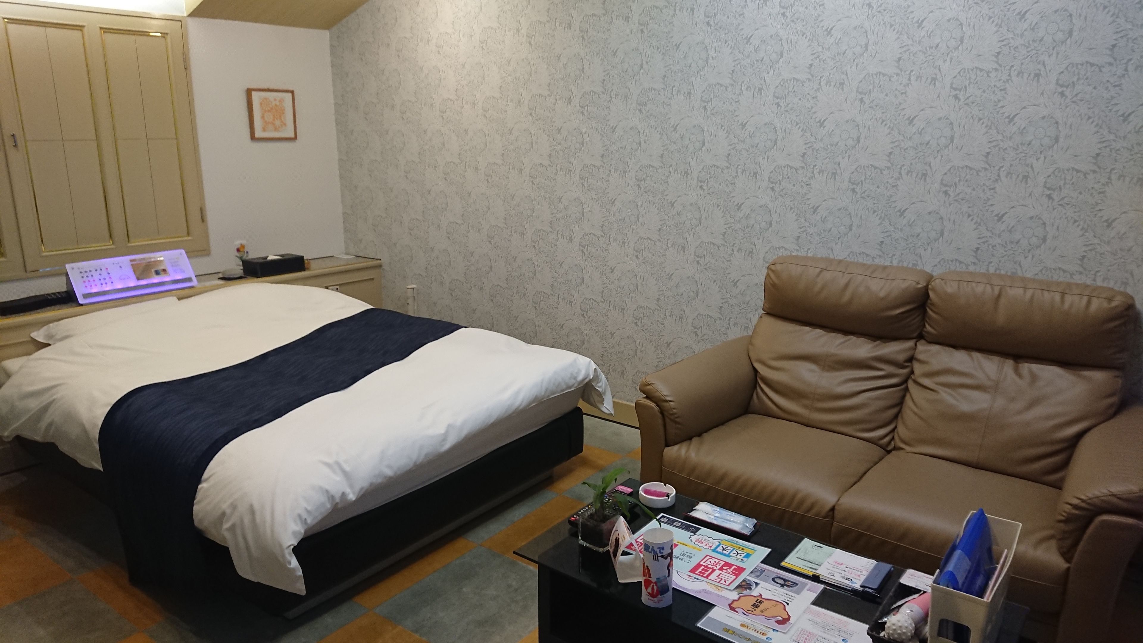 Ein Hotelzimmer mit einem Bett, einem braunen Ledersofa, einem Couchtisch mit verschiedenen Gegenständen, einem Nachttisch und einer wandmontierten Steuerung.