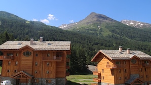 Una cabaña de madera con varios balcones y cimientos de piedra, con una cadena montañosa boscosa de fondo.