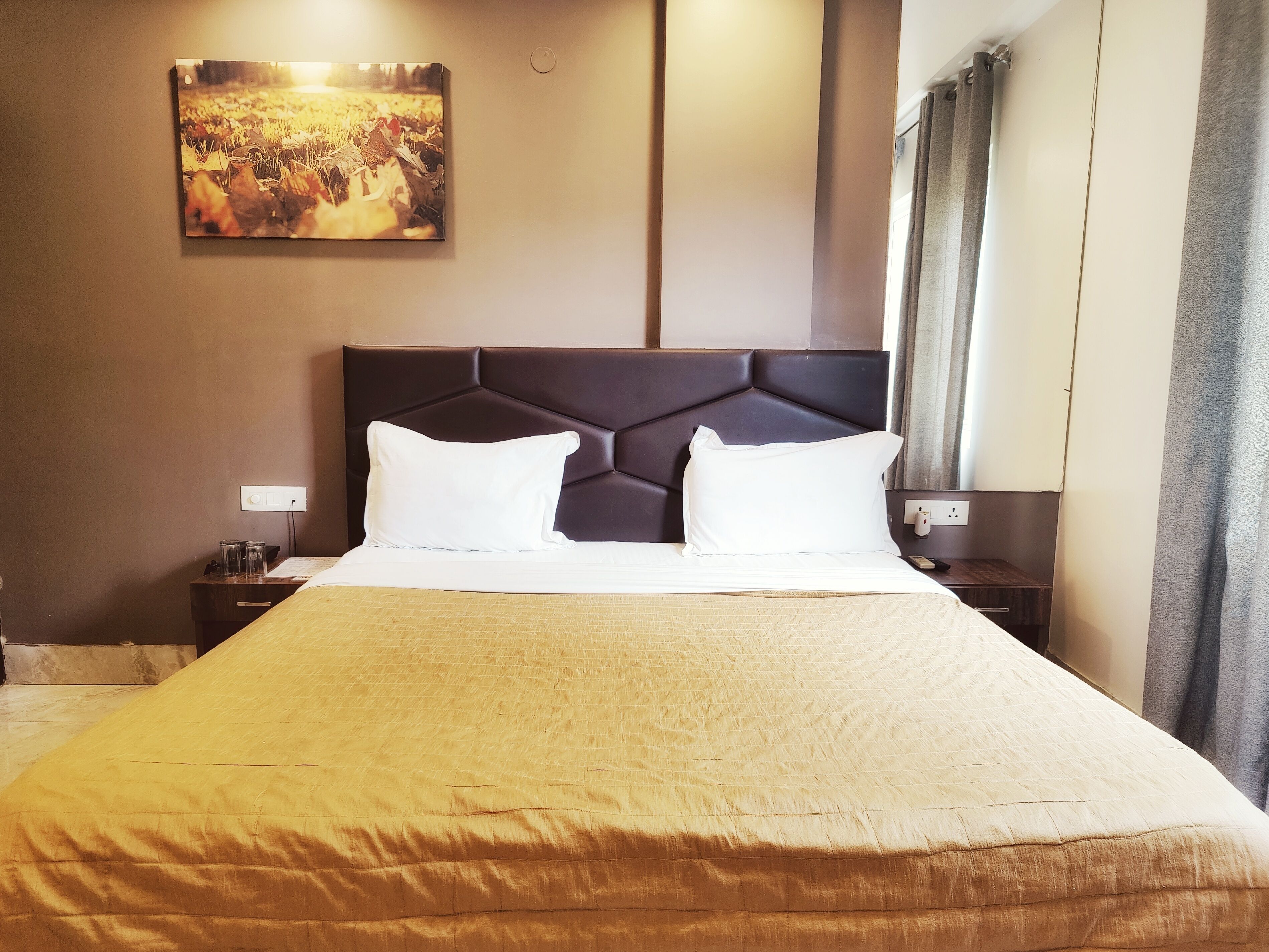 A hotel room with a large bed, two white pillows, a dark headboard, a nightstand with a lamp, and a painting on the wall.