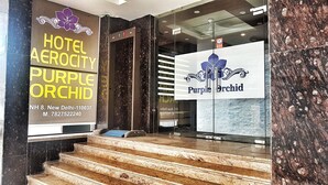 Ein Hotel Eingang mit Stufen, die zu einer Rezeption führen. Zwei auffällige Schilder sind sichtbar, eines für 'Hotel Aerocity Purple Orchid' und eines für 'Purple Orchid'.