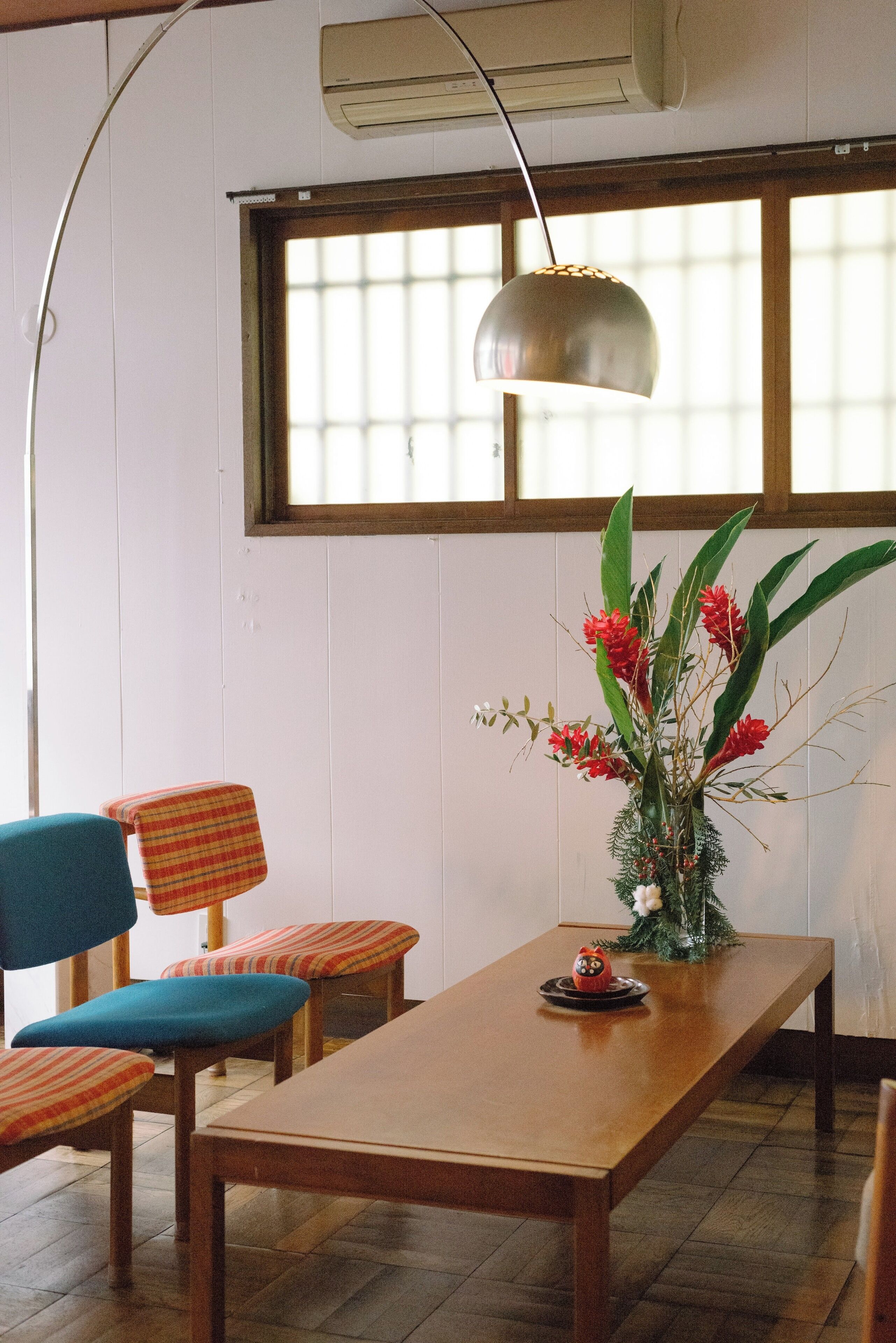 Een eetruimte met een houten tafel, blauw-rode gestreepte stoelen, een decoratieve vaas met bloemen en een hanglamp.