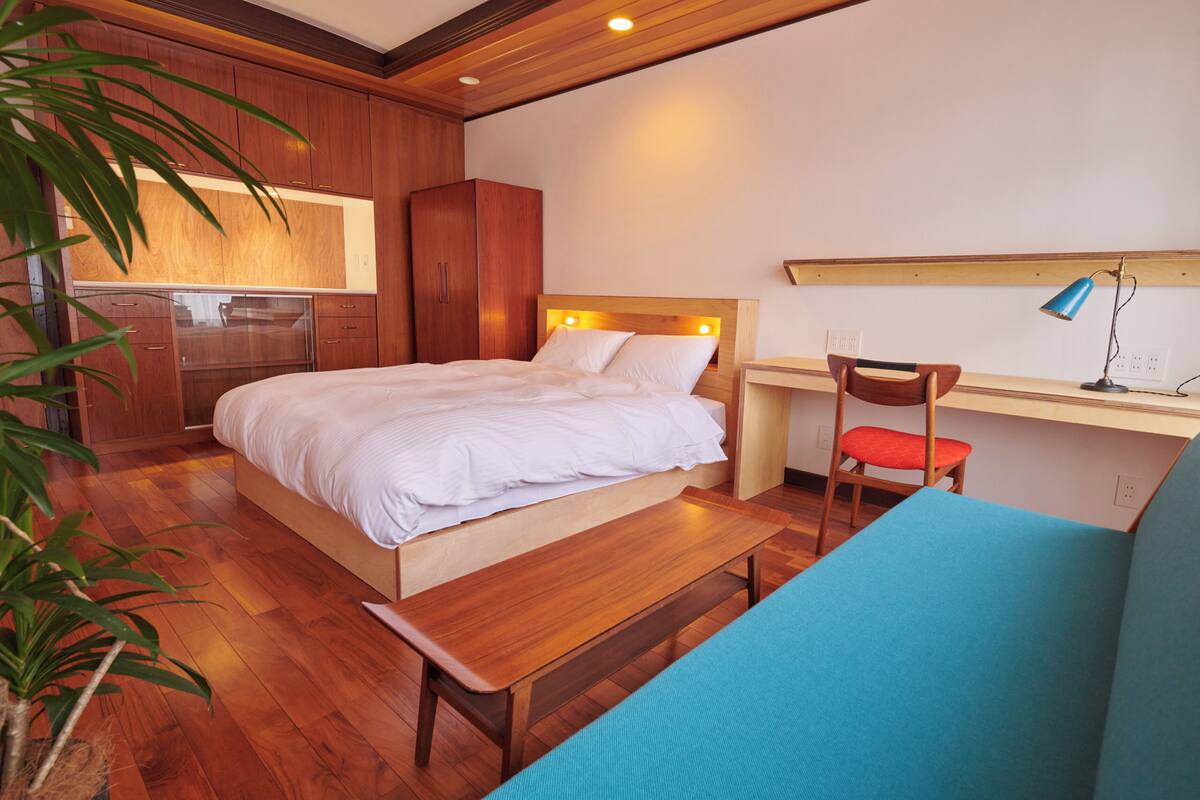 Een slaapkamer met een houten bed, een bureau met stoel, een bankje en een kamerplant.
