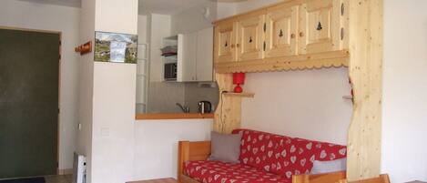A compact living space with a kitchenette, a sofa with a red heart-patterned cover, a wooden table, and a dining table with a patterned tablecloth.