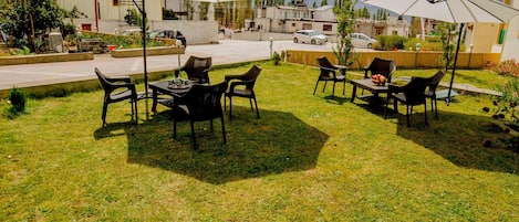 An outdoor seating area with tables and chairs under umbrellas, surrounded by a grassy lawn and a building in the background.