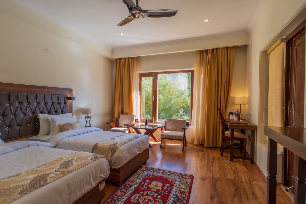A hotel room with two beds, a desk, a chair, and a view of the outdoors.