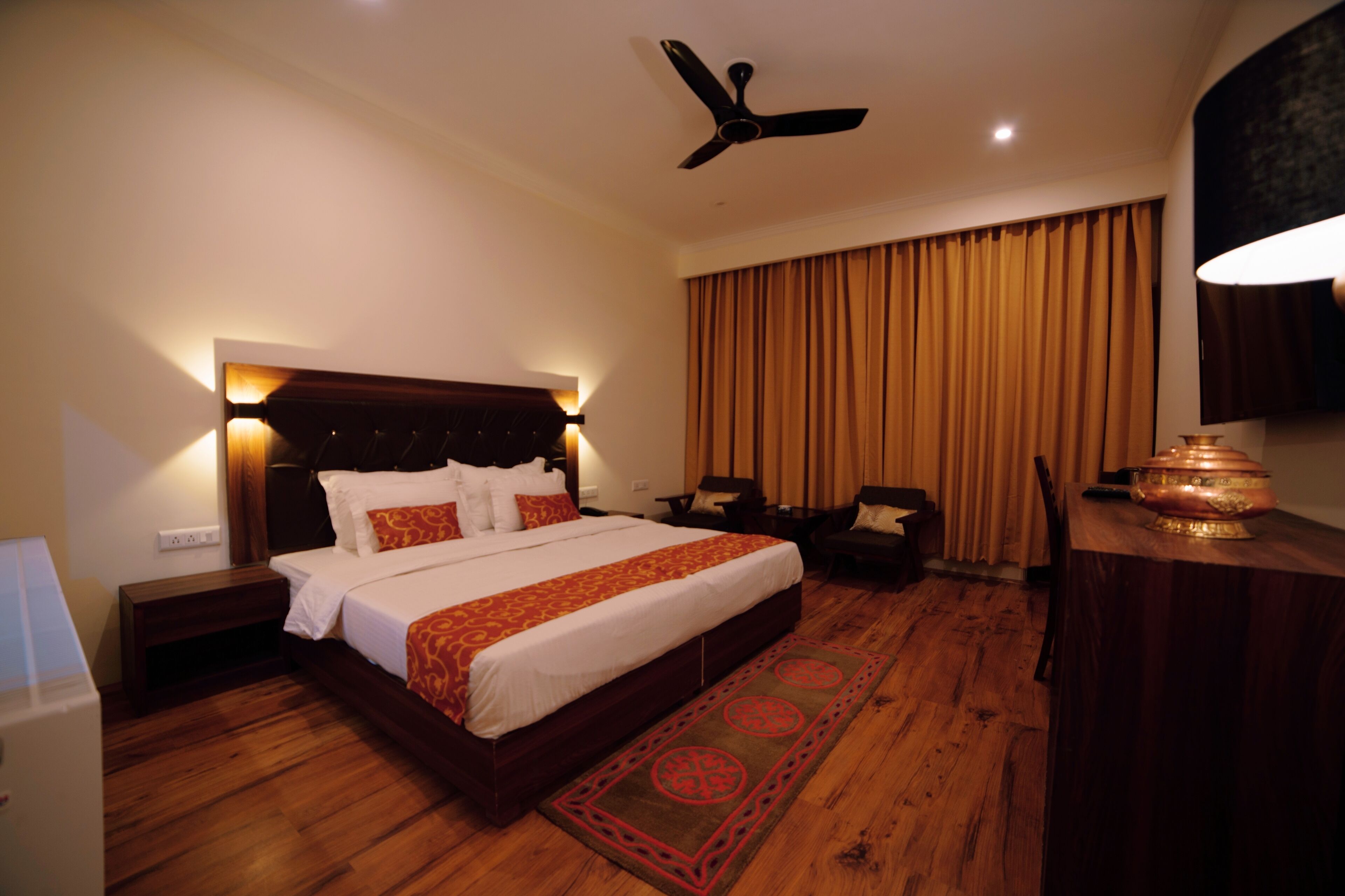 Quarto de hotel com uma cama grande, piso de madeira, ventilador de teto, mesa de cabeceira, escrivaninha com abajur e janela com cortinas.