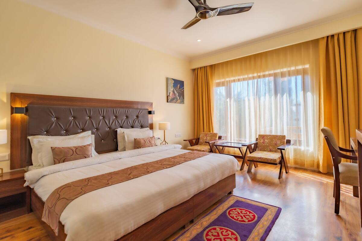 A hotel room with a large bed, a desk, two chairs, a ceiling fan, and a large window with curtains.