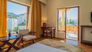 A hotel room with a bed, a desk, a chair, and a view of the city and mountains through large windows. - Hotel Leh Plaza (Leh)