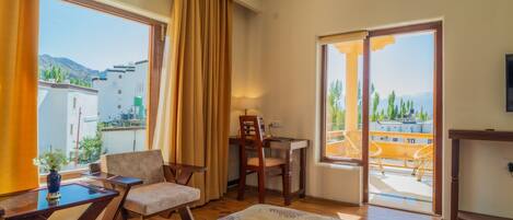 A hotel room with a bed, a desk, a chair, and a view of the city and mountains through large windows.