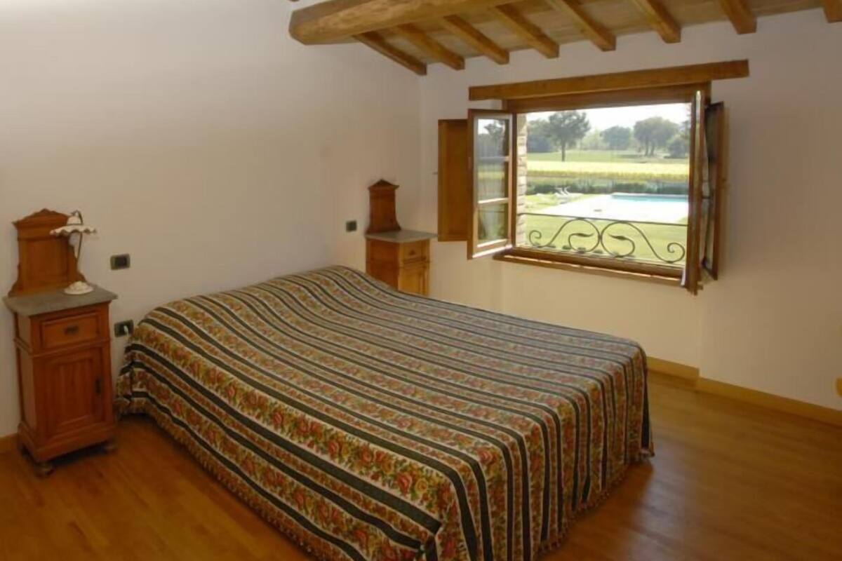 A bedroom with a wooden bed, a nightstand, a dresser, and a window with a view of a park.