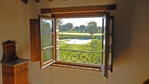 Habitació amb una finestra gran, biges de fusta al sostre i vista cap a un paisatge verd amb un edifici blanc.
