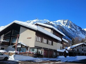 Un bâtiment dont le toit est recouvert de neige, une enseigne indiquant « L’Hôtel », et une chaîne de montagnes en arrière-plan.