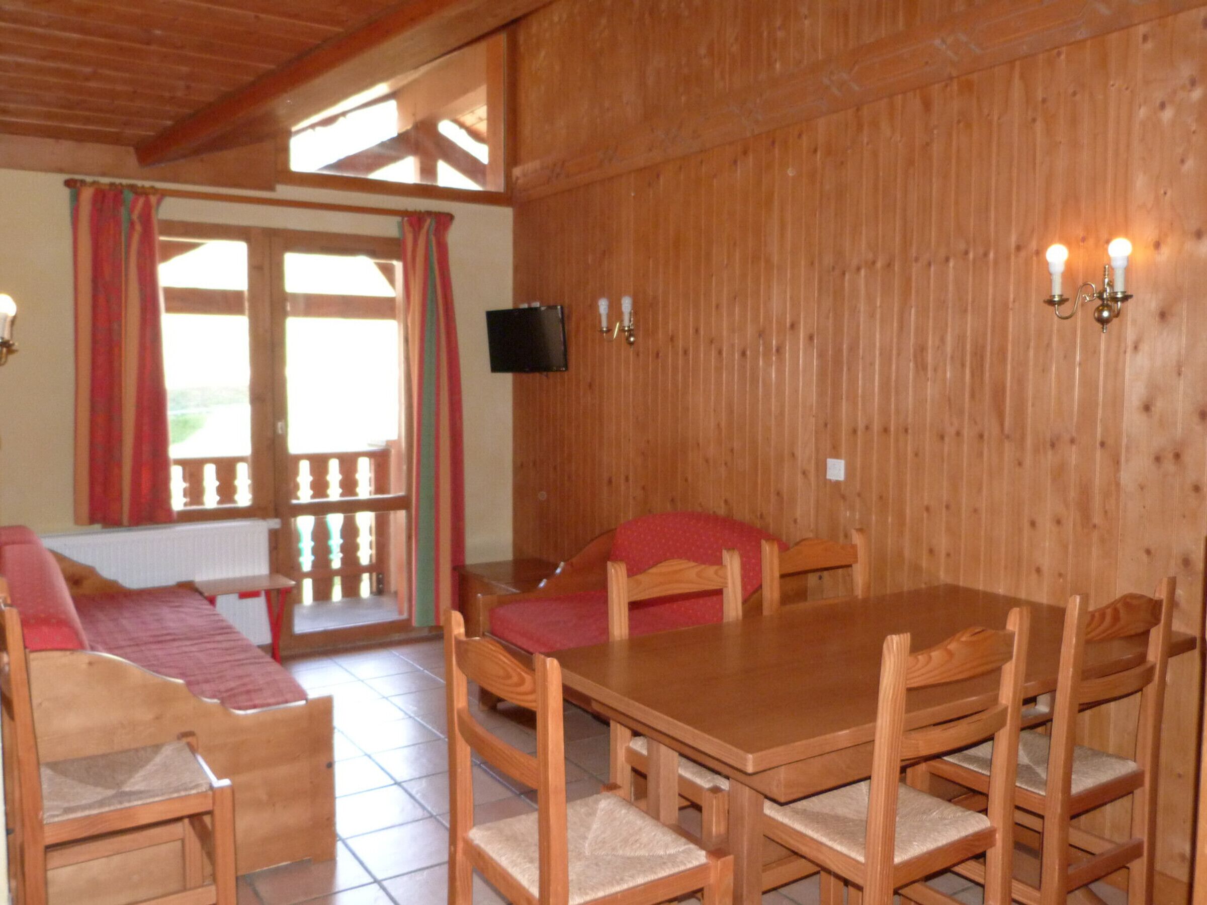 Een houten eethoek met een tafel en stoelen, een aan de muur bevestigde televisie en een raam met gordijnen.