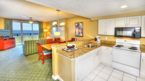 A compact kitchen with white cabinets, a microwave, and a stove. There is a dining area with a table and chairs, and a view of the ocean through large windows. - 2/2 Deluxe Ocean walk (Daytona Beach)