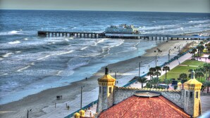 A coastal view with a pier extending into the sea, a beach with people, and a building with a red-tiled roof. - 2/2 Deluxe Ocean walk (Daytona Beach)