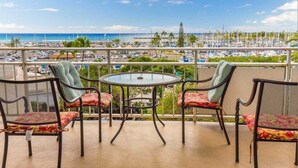 Varanda com mesa redonda e quatro cadeiras, com vista para uma marina com barcos e uma praia.