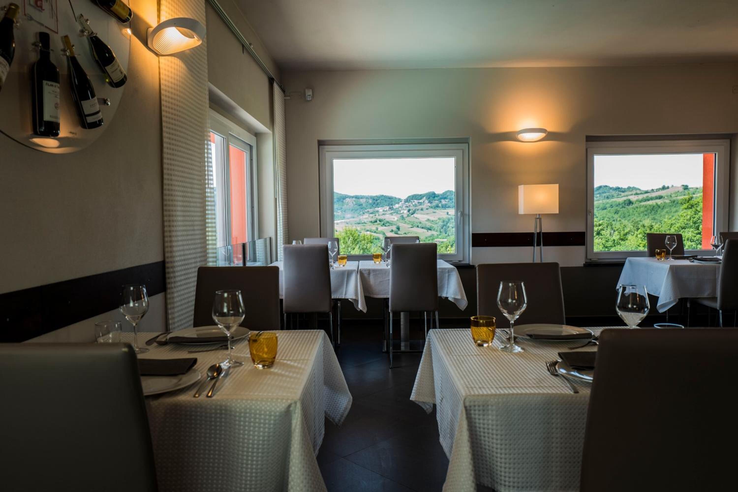 A restaurant with tables set for dining, a wine rack on the wall, and a scenic view of hills through the windows.