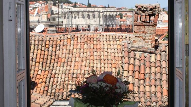 Une fenêtre ornée d’un vase de fleurs posé sur le rebord, donnant sur un paysage urbain avec des toits en tuiles rouges et des bâtiments historiques.