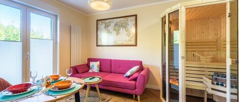 Een gezellige woonkamer met een roze bank, een eettafel gedekt met borden en glazen, en een houten sauna zichtbaar door een open deur.