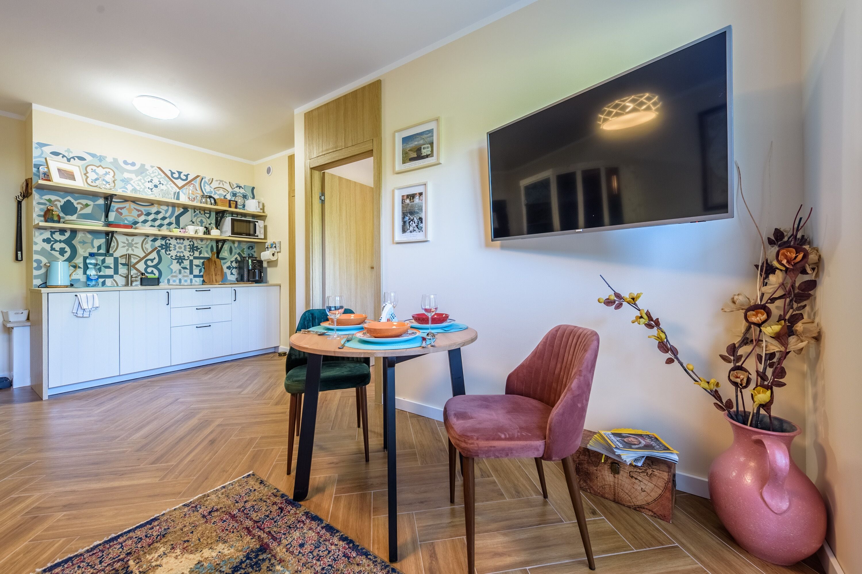 A modern kitchen with white cabinets, a dining table with chairs, and a flat-screen TV mounted on the wall.