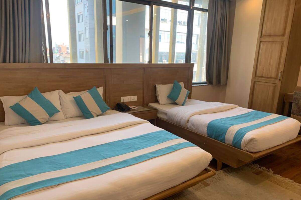 Quarto de hotel com duas camas, cada uma com cabeceira de madeira e vista da cidade através de amplas janelas.