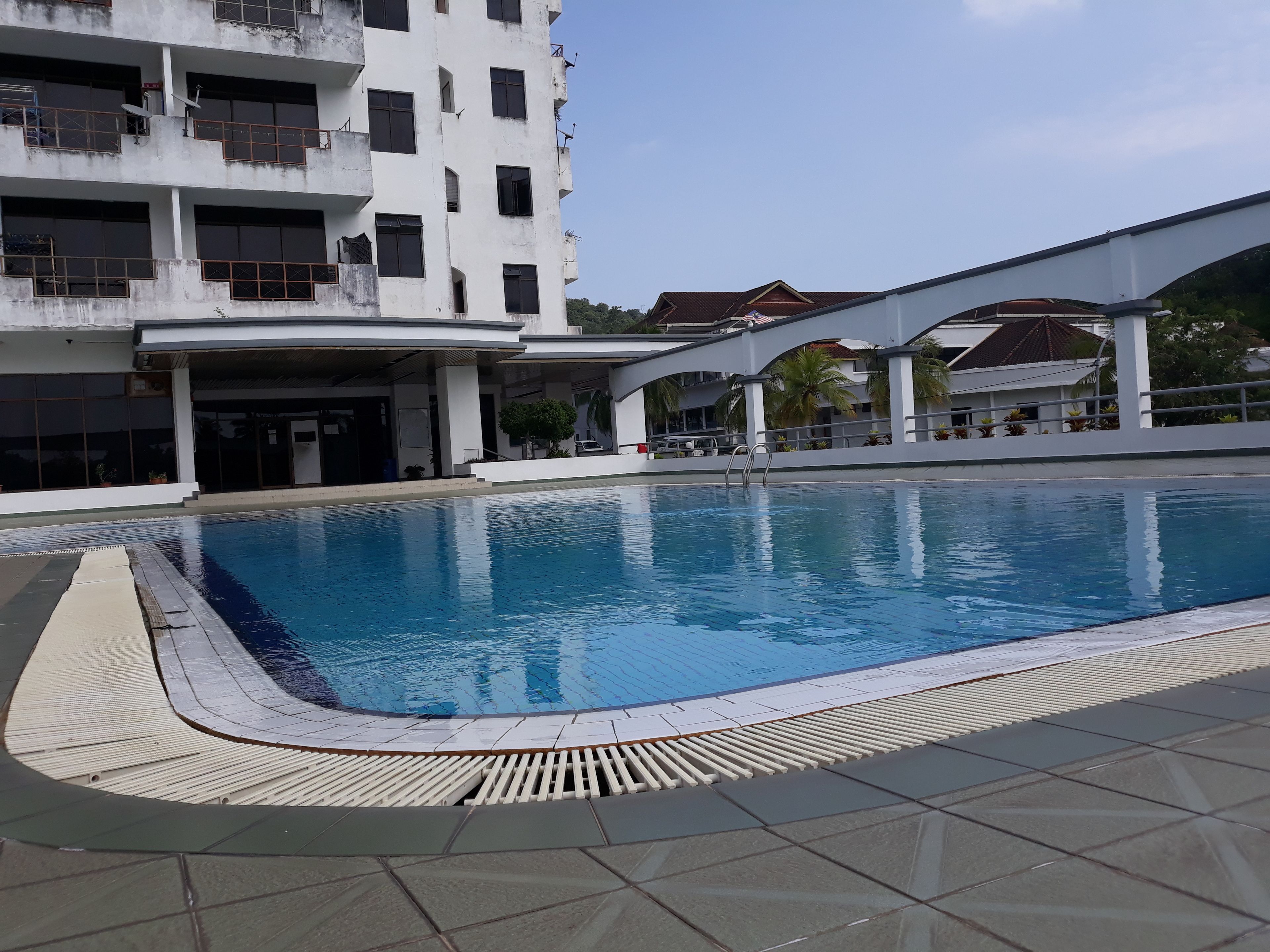 A swimming pool with a tiled deck, a covered area with a roof, and a multi-story building with balconies and satellite dishes.