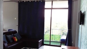 Un salon avec un canapé aux couleurs sombres, un tapis à motifs, une petite table en bois et une vue sur un jardin verdoyant, visible à travers une porte-fenêtre coulissante.