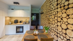 Una cocina moderna con armarios blancos, horno empotrado y cafetera. Hay una zona de comedor con mesa y sillas de madera, y una pared cubierta de leña apilada.