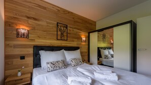 Una cama bien hecha con sábanas y toallas blancas, cabecero de madera y dos lámparas fijadas a la pared.