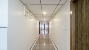 A long, narrow hallway with a polished floor, white walls, and a wooden door on the right. - Treebo Anjani Fortune Suites (Bengaluru)