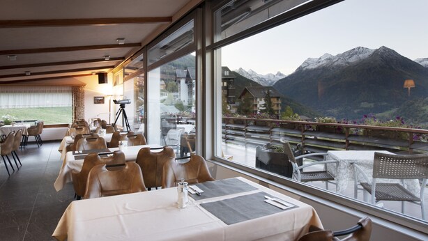 Ein Restaurant mit Tischen und Stühlen, großen Fenstern mit Blick auf die Berge und einer Terrasse mit Außenbestuhlung.