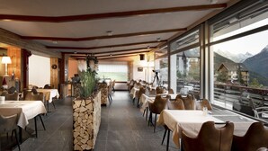 Ein Restaurant mit Tischen und Stühlen, ein Steinpflanzgefäß mit Bepflanzung und große Fenster mit Blick auf die Berge.
