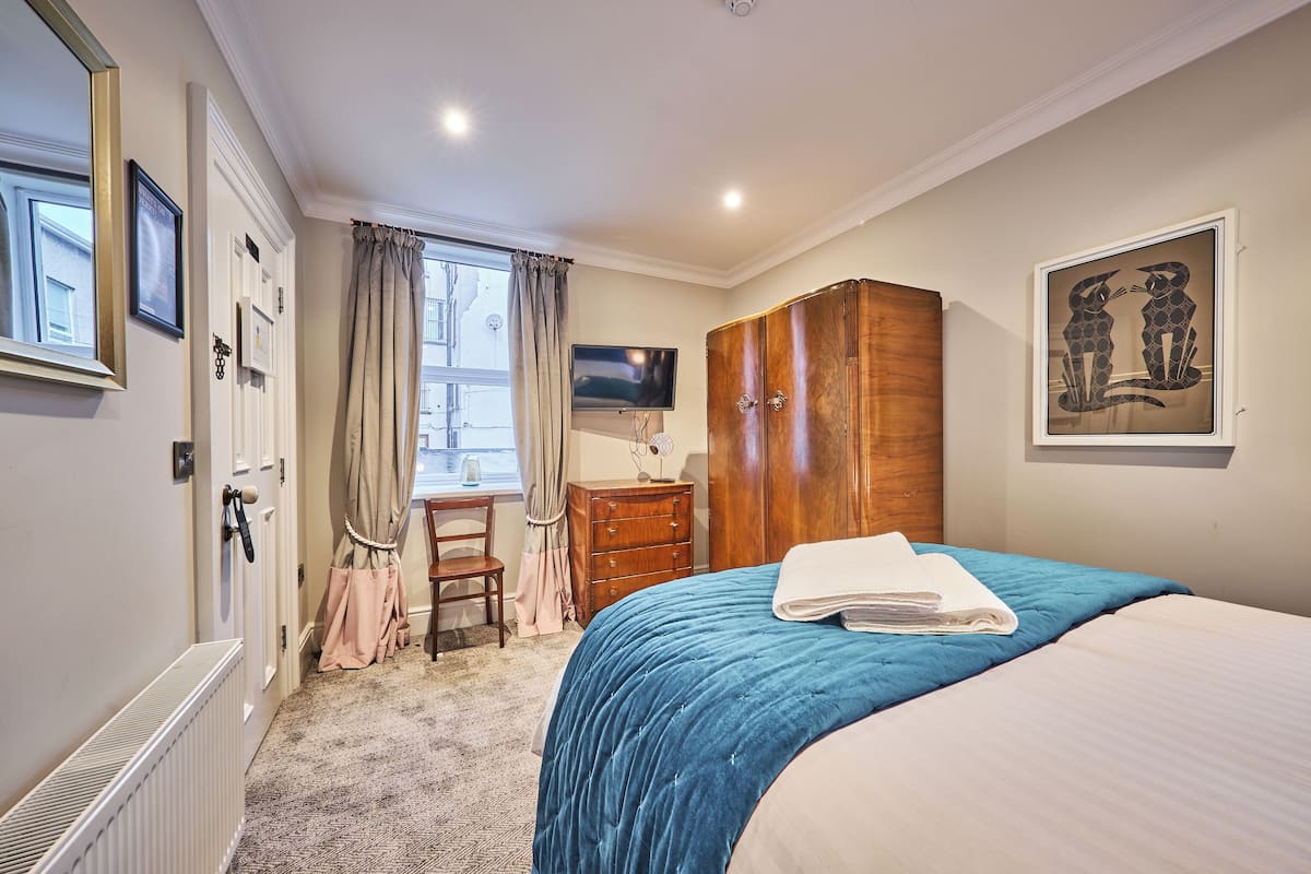 Quarto com cama, penteadeira, escrivaninha, televisão e janela com cortinas.