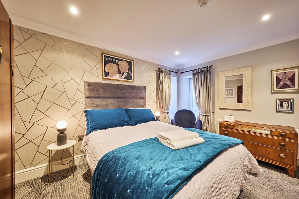 Um quarto moderno com parede estampada em padrão geométrico, uma cama grande com roupa de cama azul e branca, uma mesa de cabeceira com luminária, um aparador de madeira e um espelho na parede.