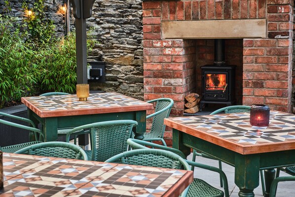 An outdoor seating area with square tables and green wicker chairs, a brick fireplace with a wood-burning stove, and a stone wall backdrop. - Art House (Londonderry)